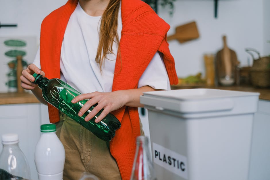 A person standing in a domestic kitchen holding a green plastic bottle with both hands, positioned near a white recycling bin labeled 'PLASTIC,' which is placed on a countertop. The individual is wear